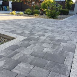 BRUK-MED Kamil Grądziel - Ułożona szara kostka brukowa w geometryczny wzór, obrzeża z jasnego grysu, mały skalniak z kamieniami i roślinami, w tle brama wjazdowa i elementy roślinności.