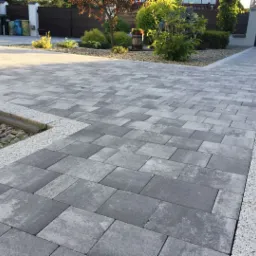 Ułożona szara kostka brukowa w geometryczny wzór, obrzeża z jasnego grysu, mały skalniak z kamieniami i roślinami, w tle brama wjazdowa i elementy roślinności.