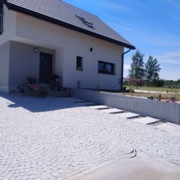 BRUK-MED Kamil Grądziel - Jasnoszara kostka granitowa ułożona w łukowy wzór na podjeździe przed nowoczesnym domem, z widocznymi pracownikami i narzędziami podczas układania.