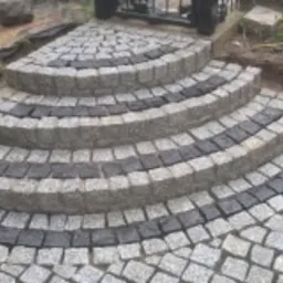 Półokrągłe schody z kostki granitowej ułożone w szaro-czarne pasy, widoczne elementy krajobrazu ogrodu w tle.