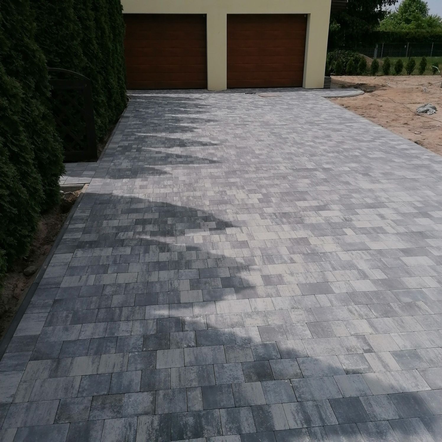 Ułożona szara kostka brukowa prowadząca do garażu z drewnianymi drzwiami, widoczne cienie drzew na powierzchni, obok żywopłot z tui.