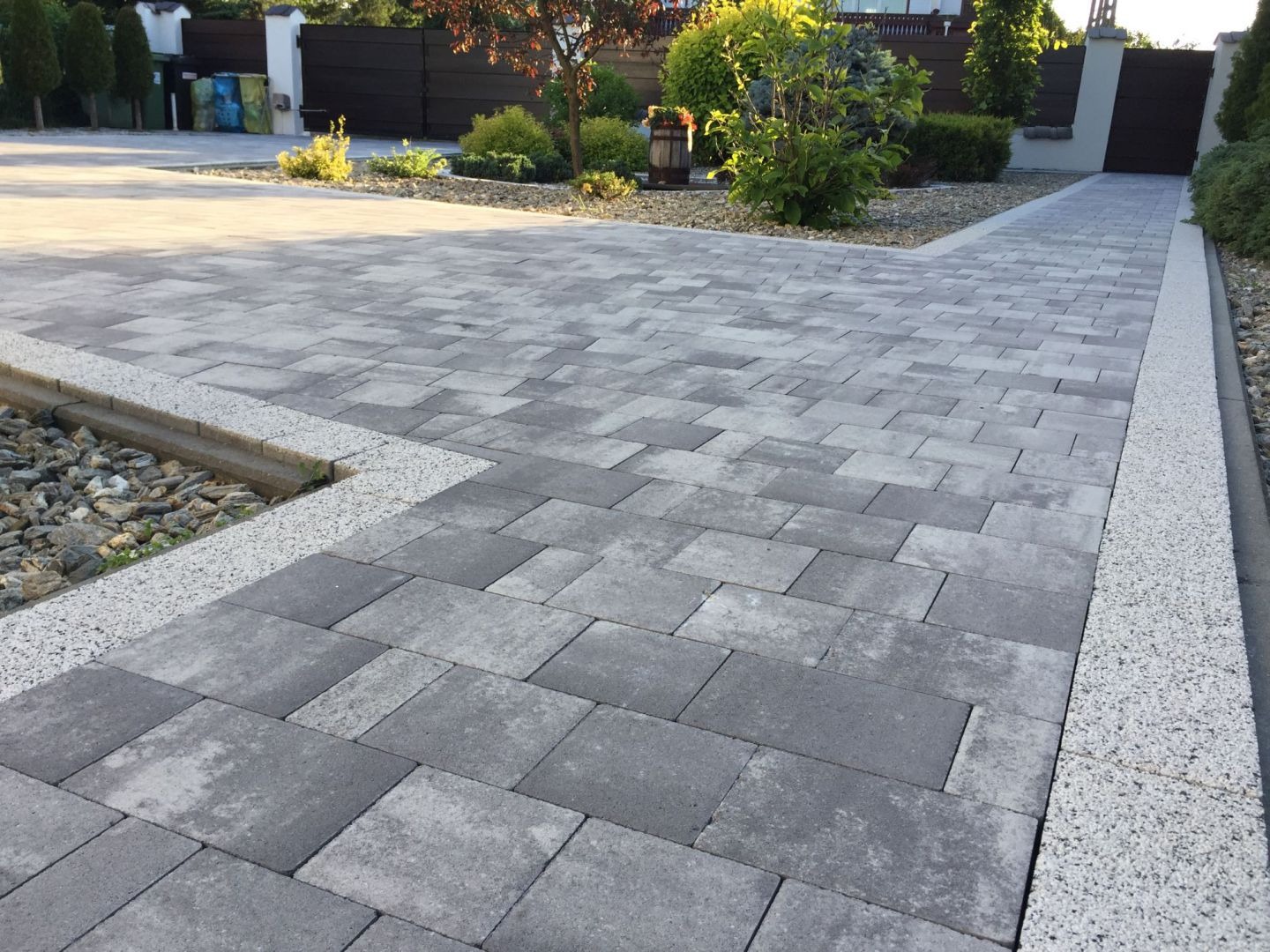 Ułożona szara kostka brukowa w geometryczny wzór, obrzeża z jasnego grysu, mały skalniak z kamieniami i roślinami, w tle brama wjazdowa i elementy roślinności.
