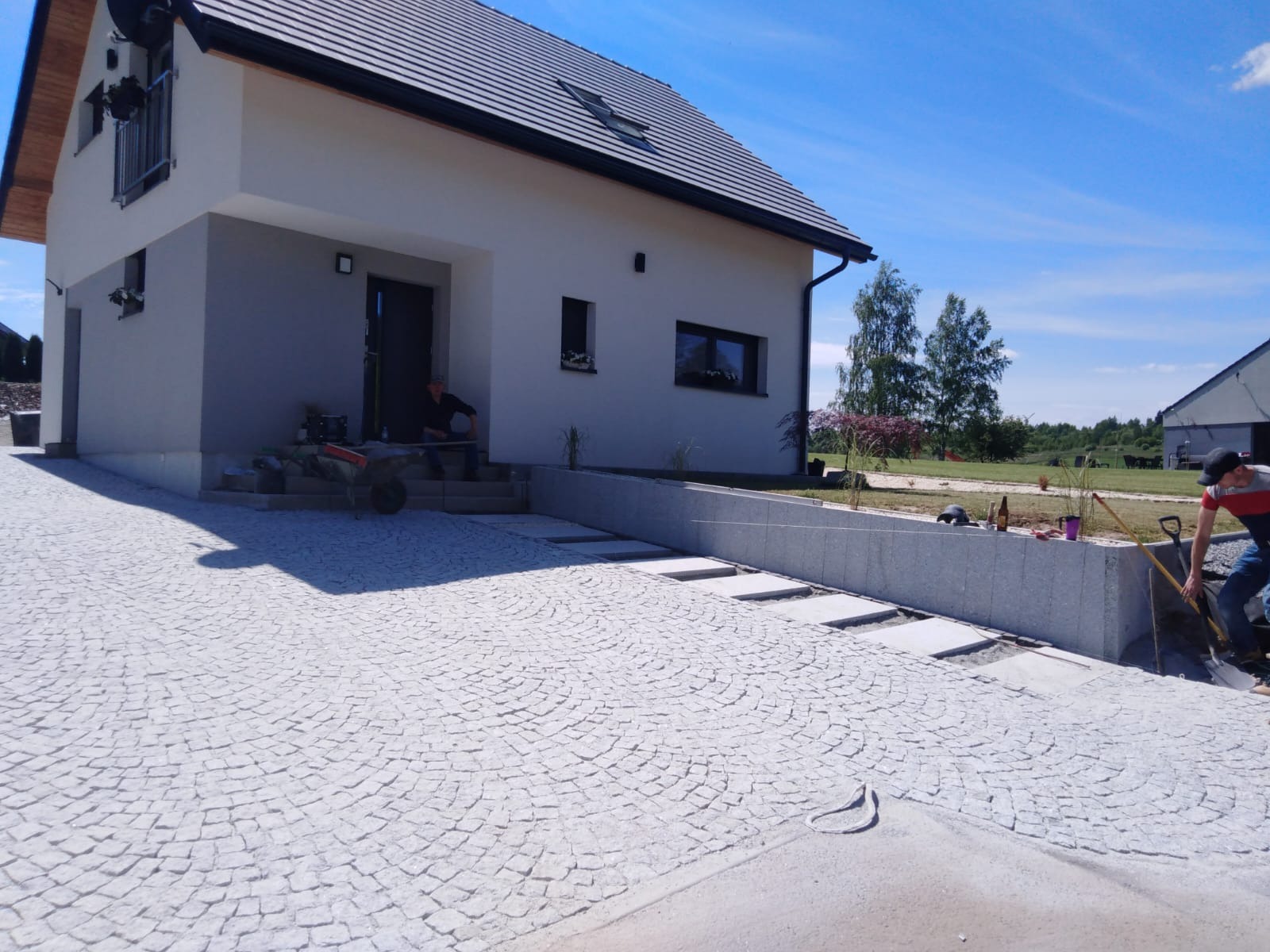 Jasnoszara kostka granitowa ułożona w łukowy wzór na podjeździe przed nowoczesnym domem, z widocznymi pracownikami i narzędziami podczas układania.