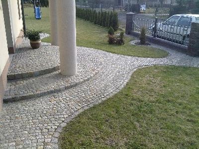 Granitowa ścieżka z kostki prowadząca od schodów domu do furtki, otoczona trawnikiem i niskimi tujami.