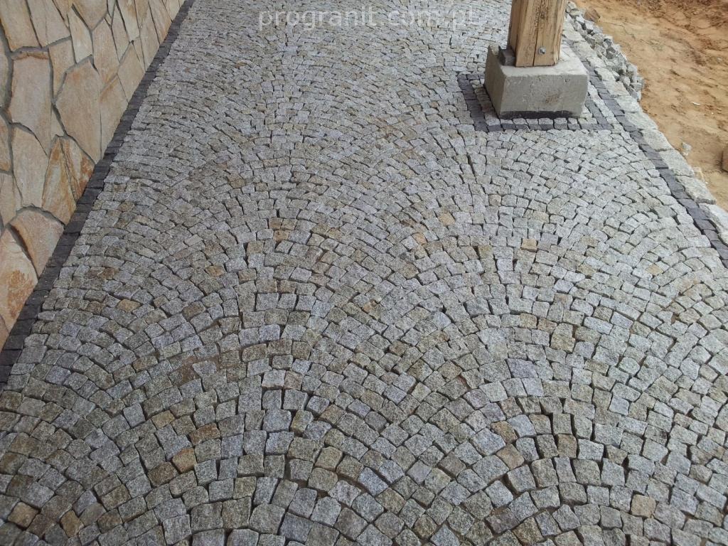 Ścieżka z szarej kostki granitowej ułożonej w wachlarzowy wzór, obok ściana z kamienia i drewniany słupek z betonową podstawą.