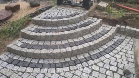 Półokrągłe schody z kostki granitowej ułożone w szaro-czarne pasy, widoczne elementy krajobrazu ogrodu w tle.