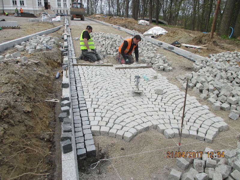 Dwóch pracowników w kamizelkach odblaskowych układa łukowy wzór z jasnoszarej kostki granitowej na podsypce piaskowej, widoczne narzędzia i stosy kostki, w tle budynek i drzewa.