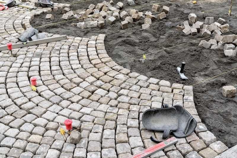 Układanie łukowatej ścieżki z kostki granitowej, widoczne narzędzia i materiały budowlane, etap prac w toku.