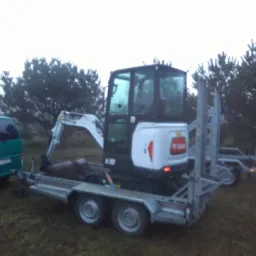 Mini koparka Bobcat na niskopodwoziowej przyczepie, holowana przez zielony samochód dostawczy na tle zalesionego terenu w pochmurny dzień.