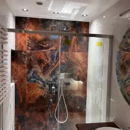 Nowoczesna kabina prysznicowa z drzwiami przesuwnymi i płytkami imitującymi marmur w odcieniach brązu i granatu, widoczny zestaw prysznicowy i umywalka.