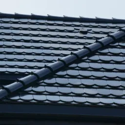 Czarna dachówka karpiówka ułożona na dachu domu jednorodzinnego. Widoczny fragment rynny i lukarna. Dach z połyskiem w słoneczny dzień.