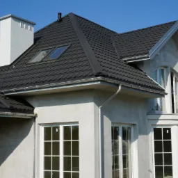 Dom z ciemnym dachem z dachówki ceramicznej, jasnymi oknami i rynnami. Widoczny komin i okna dachowe. Ściany otynkowane, przygotowane do malowania.