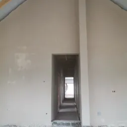 Wnętrze domu w trakcie wykańczania: świeżo otynkowane ściany w odcieniach beżu, widoczne drewniane elementy konstrukcji dachu, a w głębi korytarza postać w białym kombinezonie.