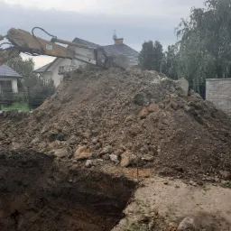 Żółta koparka nabiera ziemię z głębokiego wykopu, z tyłu widoczne domy i ogrodzenie z bloczków betonowych.