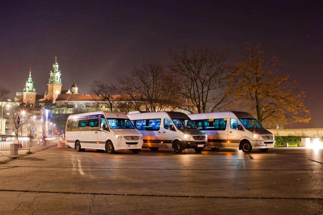 Trzy białe busy Mercedes-Benz Sprinter ustawione w rzędzie na tle nocnego krajobrazu Krakowa z Wawelem, oświetlone reflektorami i światłami ulicznymi.