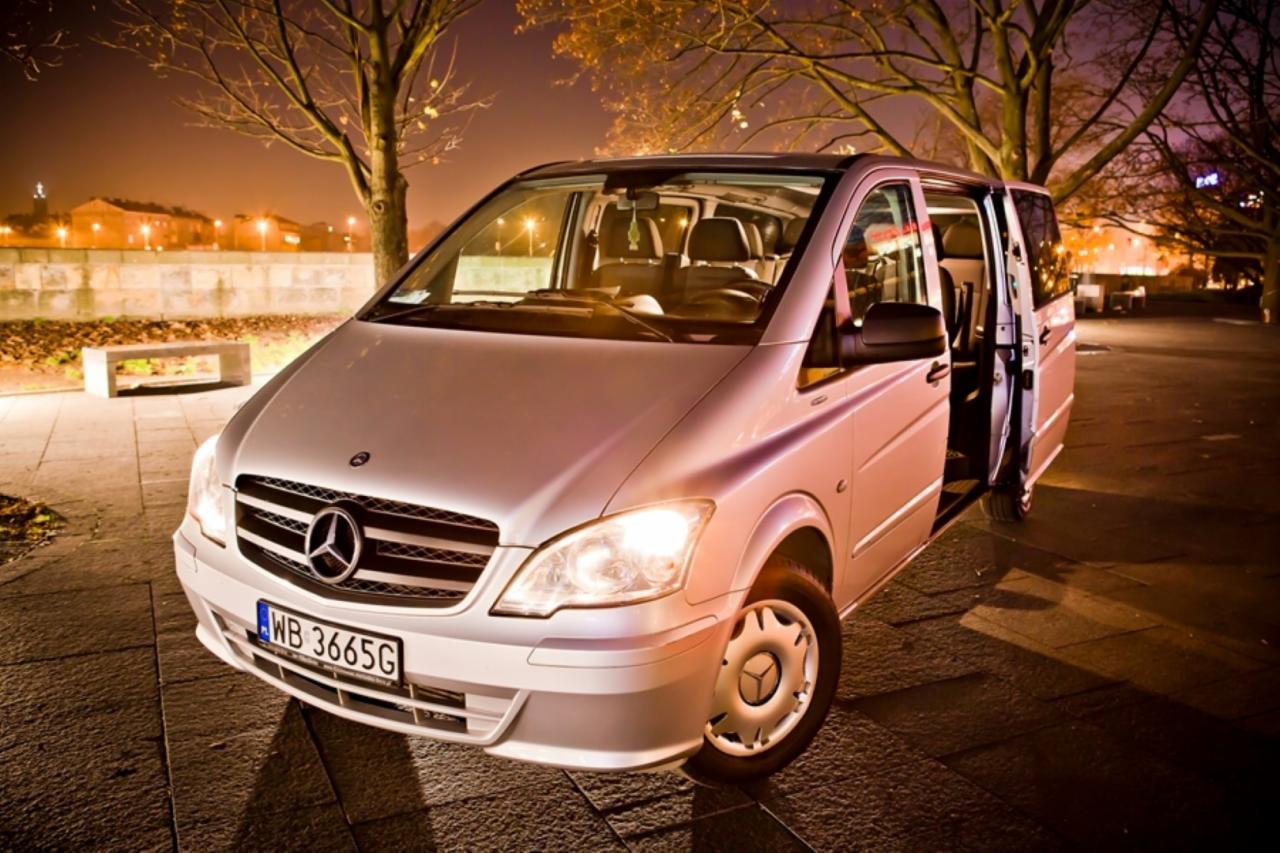 Srebrny Mercedes-Benz Vito z otwartymi drzwiami przesuwnymi, zaparkowany na tle nocnego miejskiego parku z oświetlonymi budynkami w oddali.