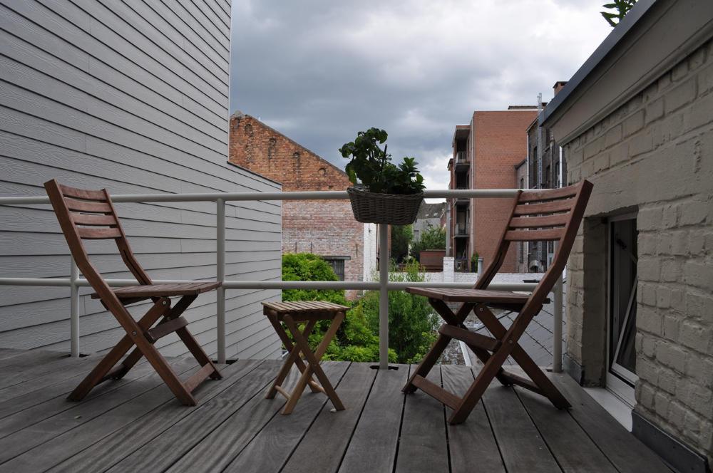 Drewniany taras z dwoma składanymi krzesłami i małym stolikiem, donica z zielonymi roślinami wisząca na białej balustradzie, szare deski podłogowe, budynki w tle pod pochmurnym niebem.