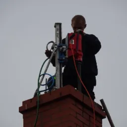 Pracownik na szczycie ceglanego komina, używa wiertnicy z wiertłem koronowym do wiercenia otworów. Widoczne przewody i elementy mocujące. Szare niebo w tle.
