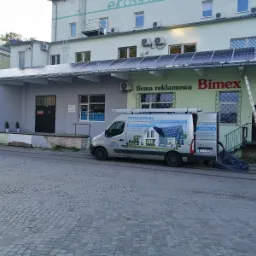 Instalacja paneli fotowoltaicznych na dachu budynku firmy reklamowej Bimex, widoczny samochód dostawczy firmy fotowoltaicznej Pertis z reklamą na karoserii oraz drabina oparta o budynek.