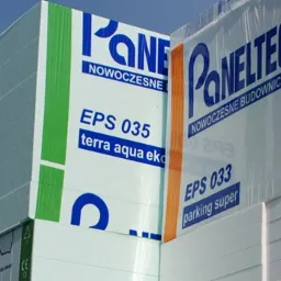 Pakiety materiałów izolacyjnych Paneltech, w tym białe płyty EPS 033 i EPS 035 oraz szare płyty grafitowe, składowane na zewnątrz pod błękitnym niebem.