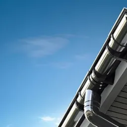 Szczegółowe ujęcie narożnika dachu z czarnymi rynnami, widoczne panele podbitki w odcieniach szarości oraz intensywne błękitne niebo w tle.