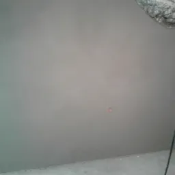 Świeżo otynkowana szara ściana z widocznym otworem na gniazdko elektryczne i fragmentem surowego muru w rogu, ukazująca etap prac wykończeniowych.