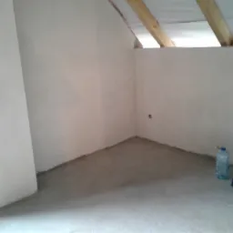 Poddasze po wylewce, widoczne drewniane belki stropowe, świeżo wylana posadzka, butelka wody i torba robocza w rogu.