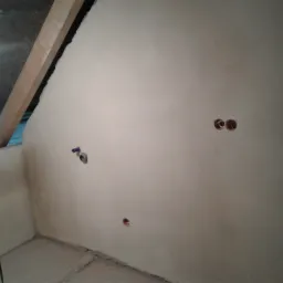 Wnętrze w trakcie prac wykończeniowych: gładka, jasna ściana z widocznymi otworami na instalacje, surowy strop z drewnianą belką i fragmentem niebieskiej folii, oraz fragment posadzki.