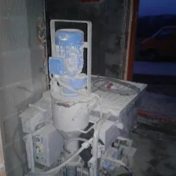 Agregat tynkarski, pokryty pyłem, stoi w surowym wnętrzu budynku w trakcie prac wykończeniowych, z widocznym fragmentem czerwonego samochodu na zewnątrz.