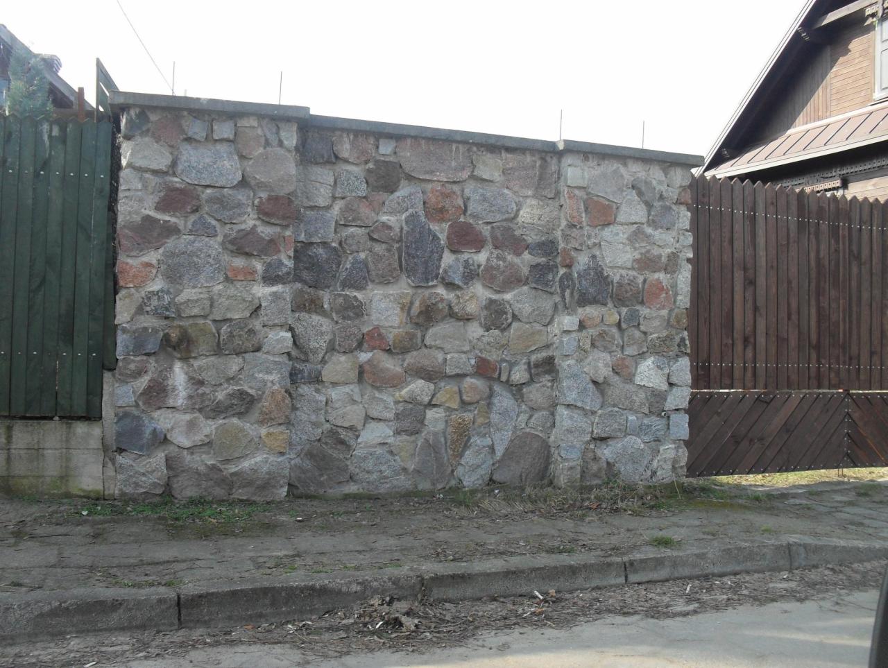 Kamienny mur ogrodzeniowy z nieregularnych, szarych i brązowych kamieni, po lewej stronie zielone drewniane ogrodzenie, po prawej brązowa drewniana brama, widoczne elementy domu w tle.