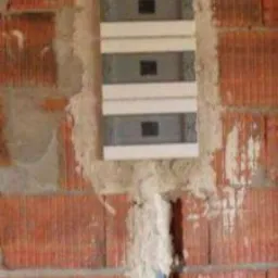 Niewykończona ściana z czerwonej cegły z osadzoną, białą skrzynką rozdzielczą z trzema bezpiecznikami i widocznymi przewodami w dolnej części.