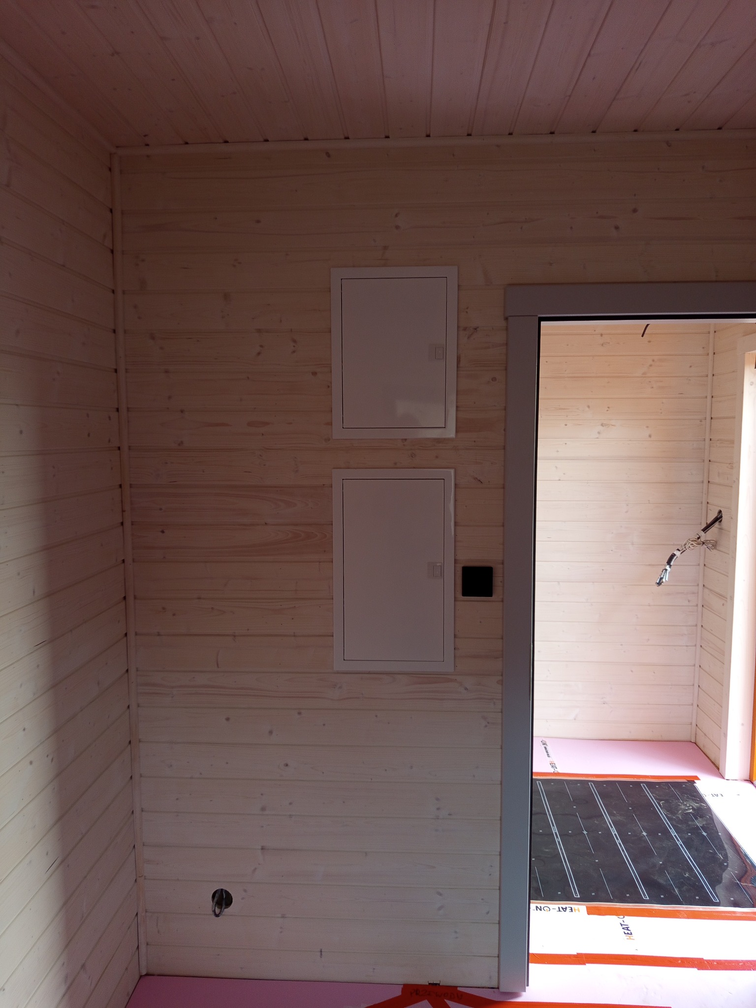 Wnętrze wykończone boazerią z dwoma białymi skrzynkami elektrycznymi na ścianie. Widoczna instalacja ogrzewania podłogowego na różowej izolacji.