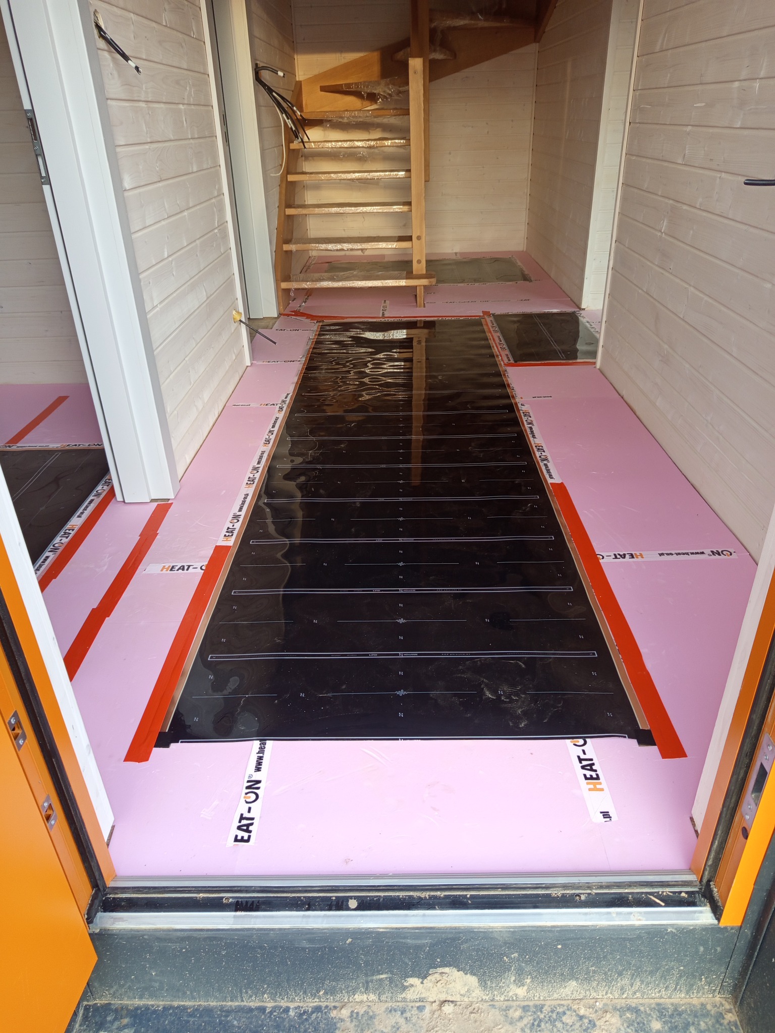 Montaż ogrzewania podłogowego: czarna mata grzewcza na różowym styropianie, widok z progu, drewniane ściany i schody w tle. Etap prac przed wylaniem posadzki.