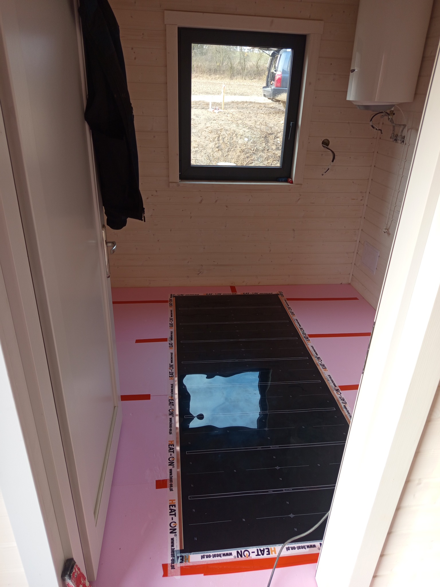 Montaż folii grzewczej HEAT-ON na różowej izolacji podłogowej w drewnianym domku. Widoczne okno i bojler na ścianie. Ujęcie zza progu.