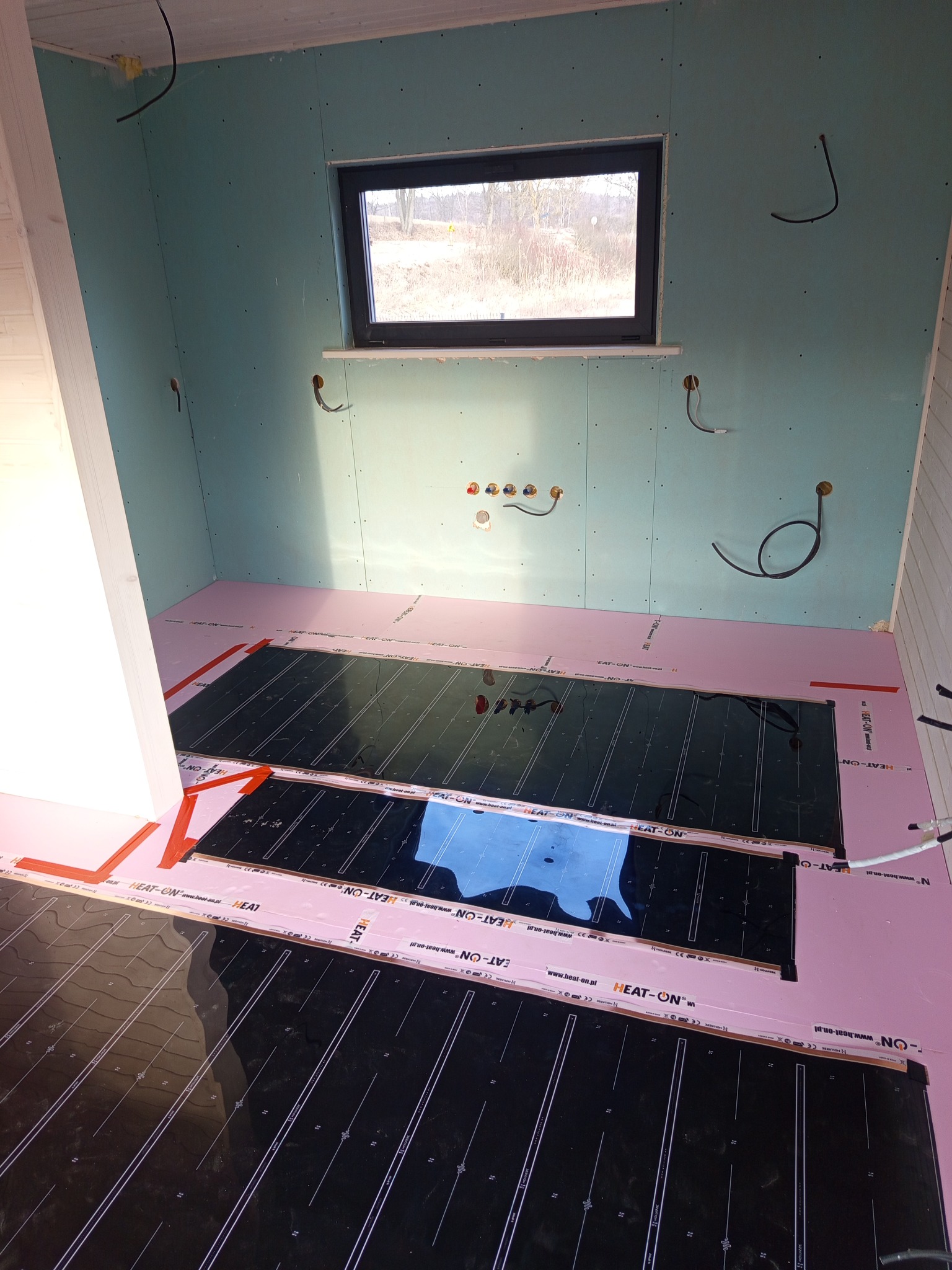 Wnętrze w trakcie prac: różowa izolacja podłogowa z czarnymi matami grzewczymi, ściany wykończone płytami gipsowo-kartonowymi, widoczne przewody elektryczne.