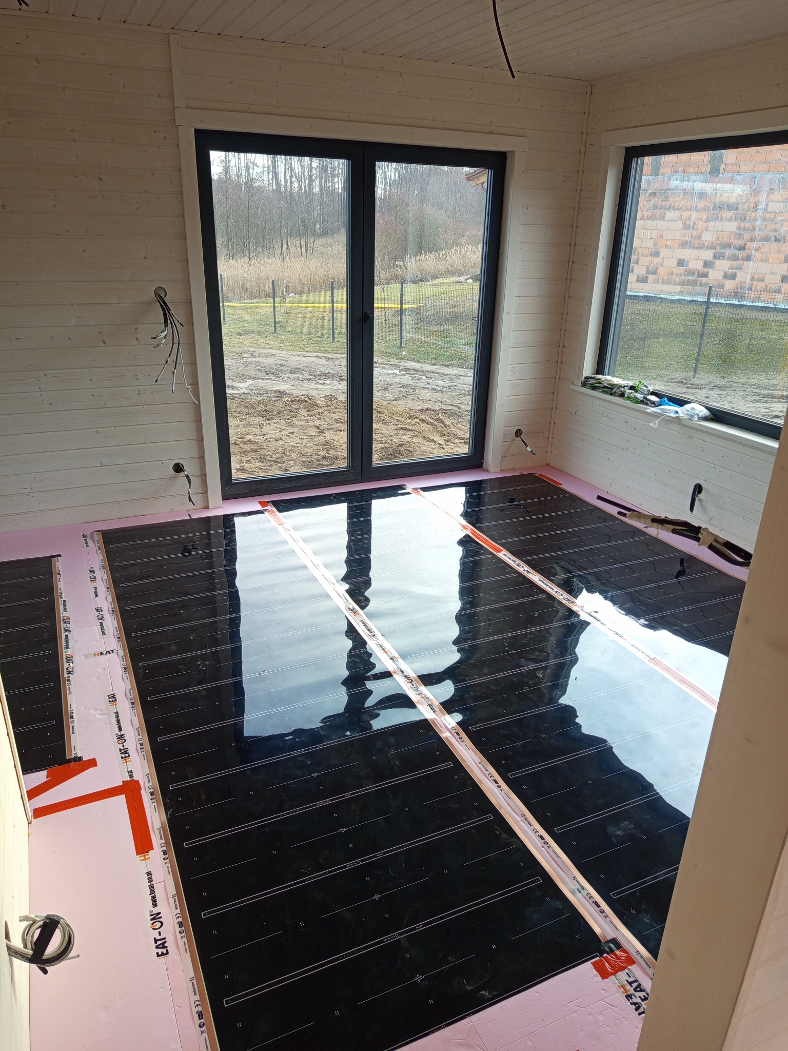 Montaż folii grzewczej na podłodze w drewnianym domu. Widoczne czarne maty grzewcze na różowym podkładzie izolacyjnym. Okna z widokiem na zewnątrz.