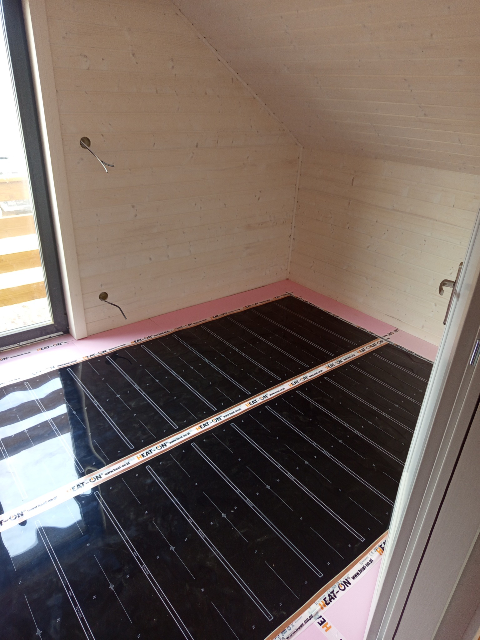 Montaż folii grzewczej HEAT-ON na podłodze w drewnianym pokoju na poddaszu. Widoczne kable elektryczne wystające ze ściany. Różowa izolacja termiczna przy ścianach.