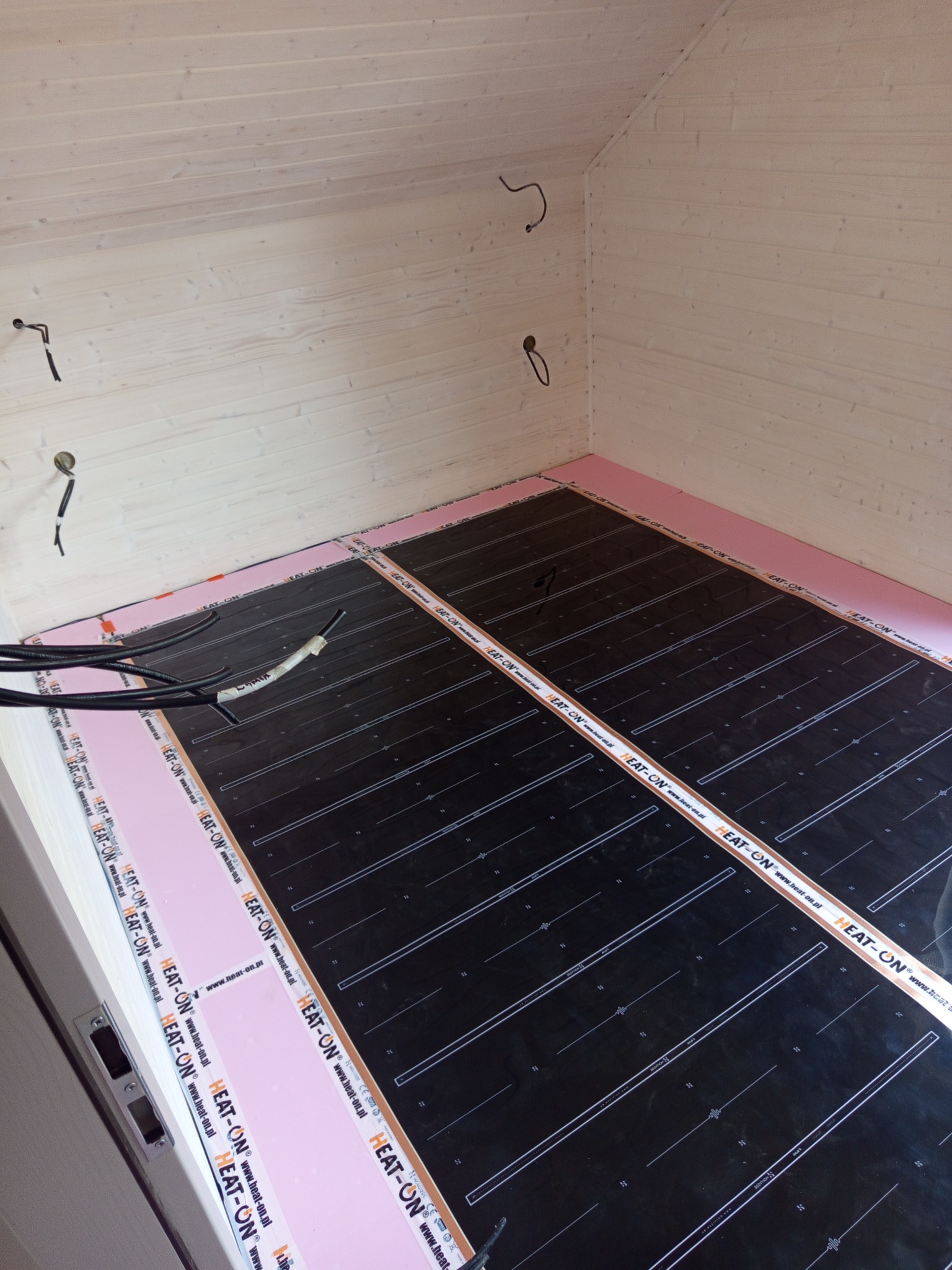 Montaż ogrzewania podłogowego: czarne maty grzewcze HEAT-ON ułożone na różowej izolacji w pomieszczeniu z boazerią. Widoczne kable elektryczne.