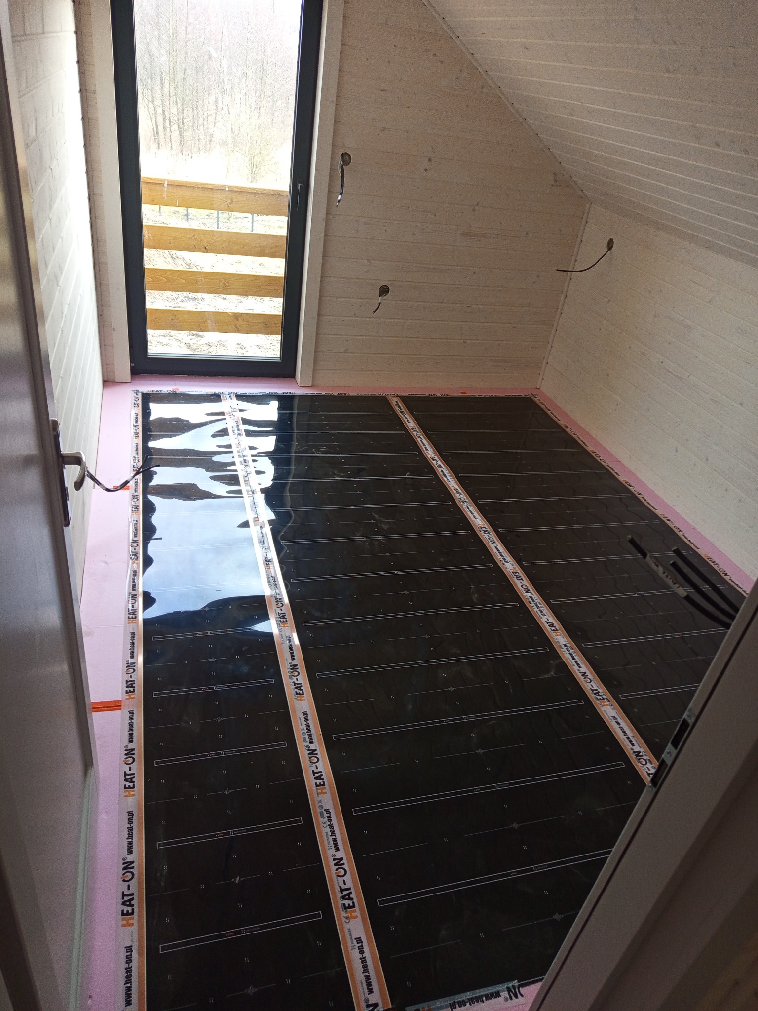 Montaż folii grzewczej EAT-ON na podłodze poddasza z drewnianą boazerią, widok z góry na rozłożoną instalację przed wylaniem wylewki. Widoczne okno dachowe i przewody.