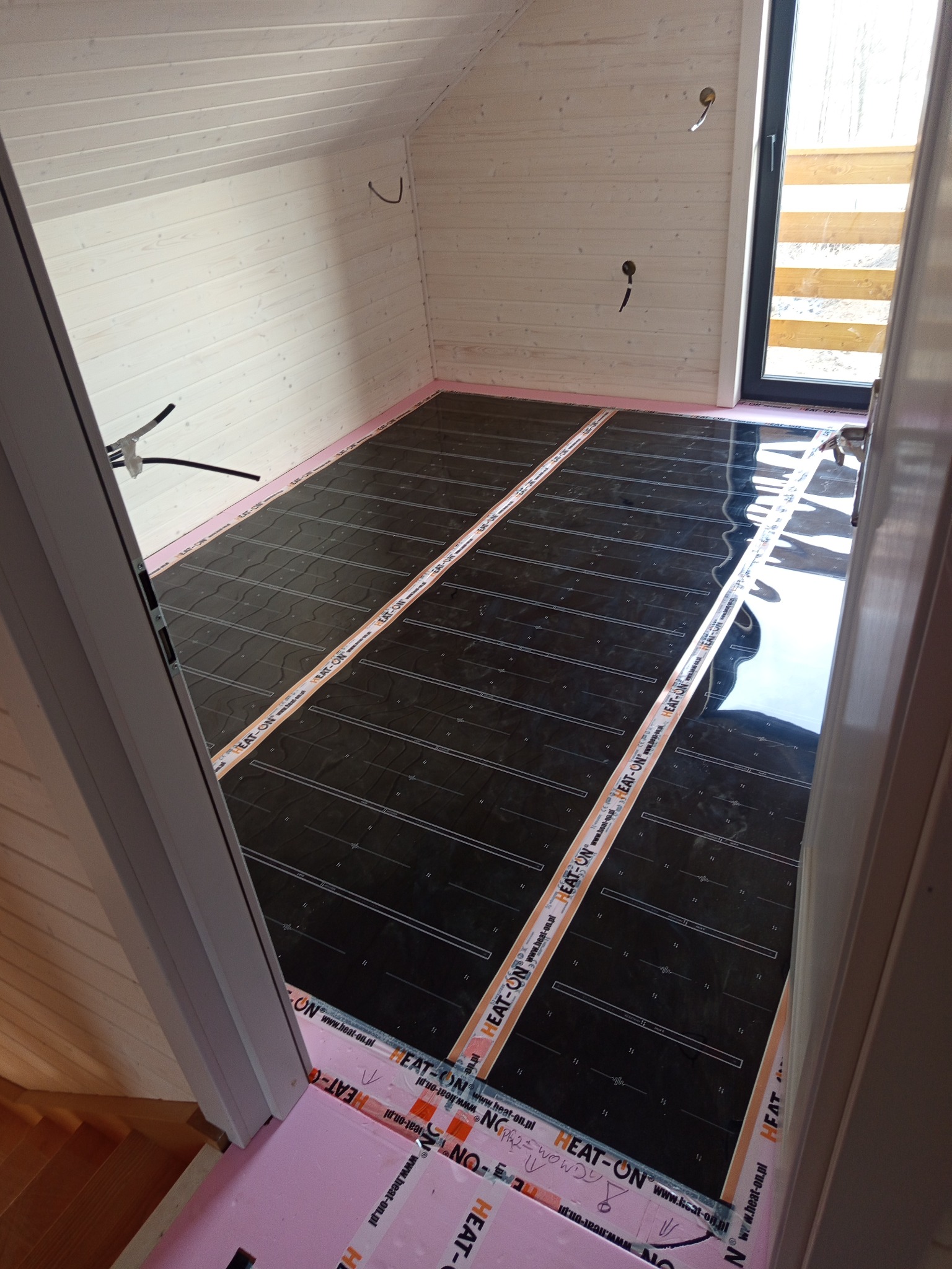 Poddasze z ułożoną folią grzewczą na podłodze, widoczne białe deski na ścianach i skosach, różowa izolacja brzegowa, czarna folia grzewcza z napisami. Widok z góry.