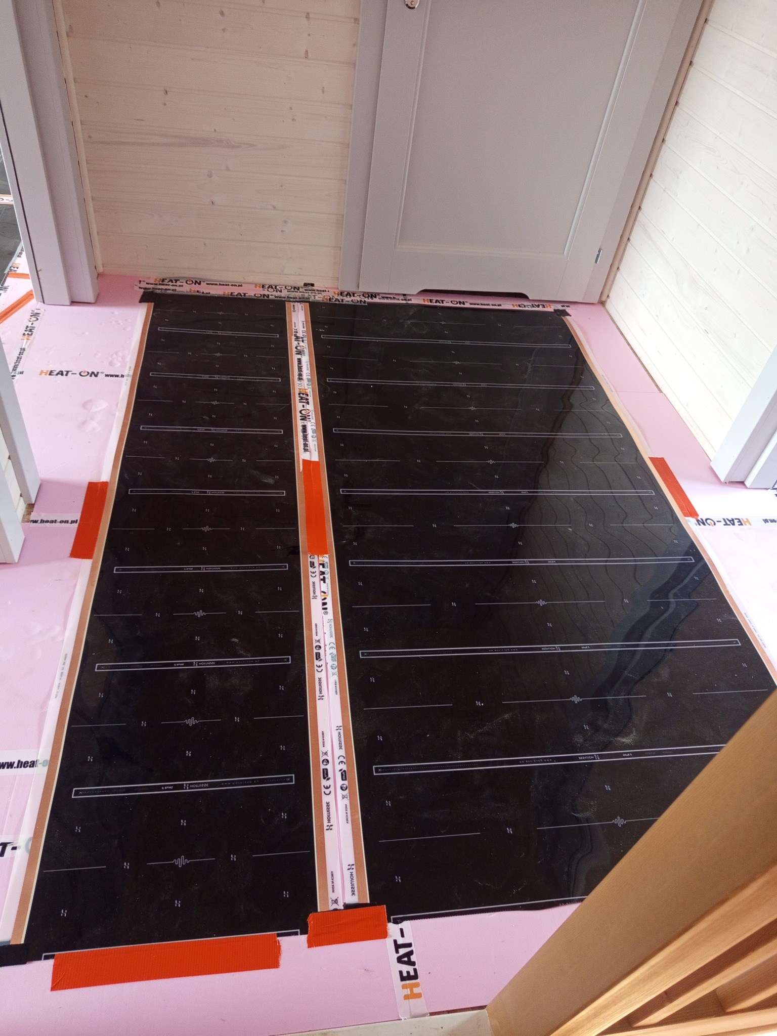 Montaż folii grzewczej HEAT-ON na różowym styropianie, widok z góry. Elementy instalacji elektrycznej pod ogrzewanie podłogowe w drewnianym wnętrzu.