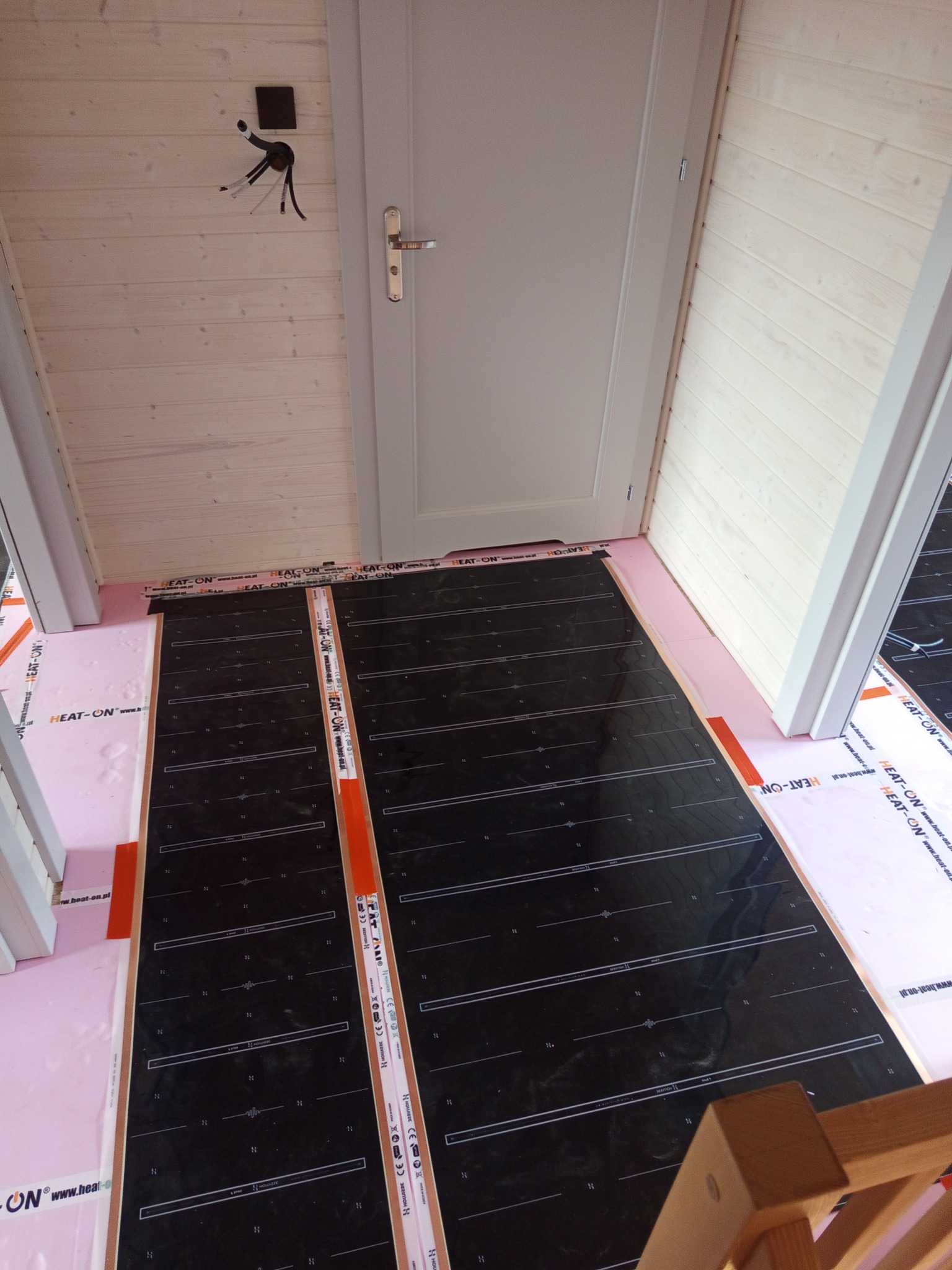 Montaż folii grzewczej HEAT-ON na różowym styropianie podłogowym w jasnym pomieszczeniu z drewnianą boazerią i białą stolarką drzwiową. Widoczne kable elektryczne.