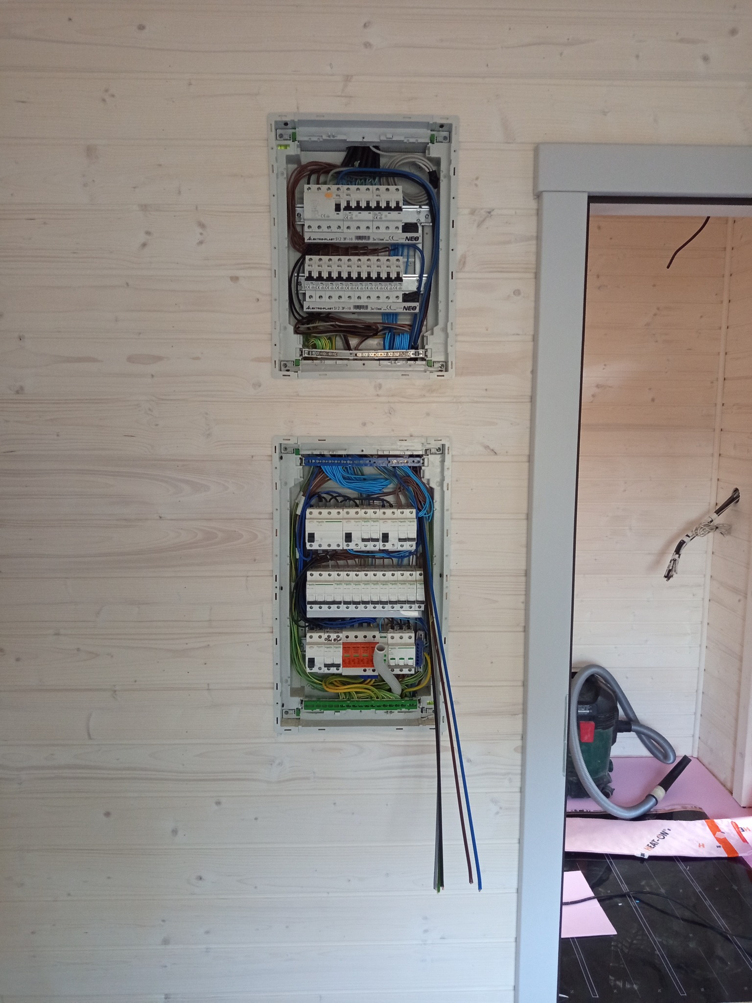 Dwie skrzynki elektryczne z zainstalowanymi bezpiecznikami na ścianie z jasnego drewna. Widoczne kable i przewody. Obok framuga drzwi i odkurzacz na podłodze z ogrzewaniem.
