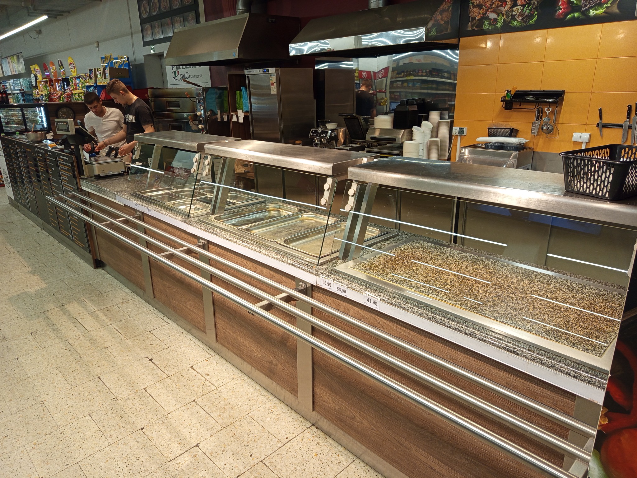 Nowoczesna lada chłodnicza z ekspozycją potraw w supermarkecie. Widoczne metalowe elementy, drewnopodobne wykończenie i pracownicy obsługujący klientów.