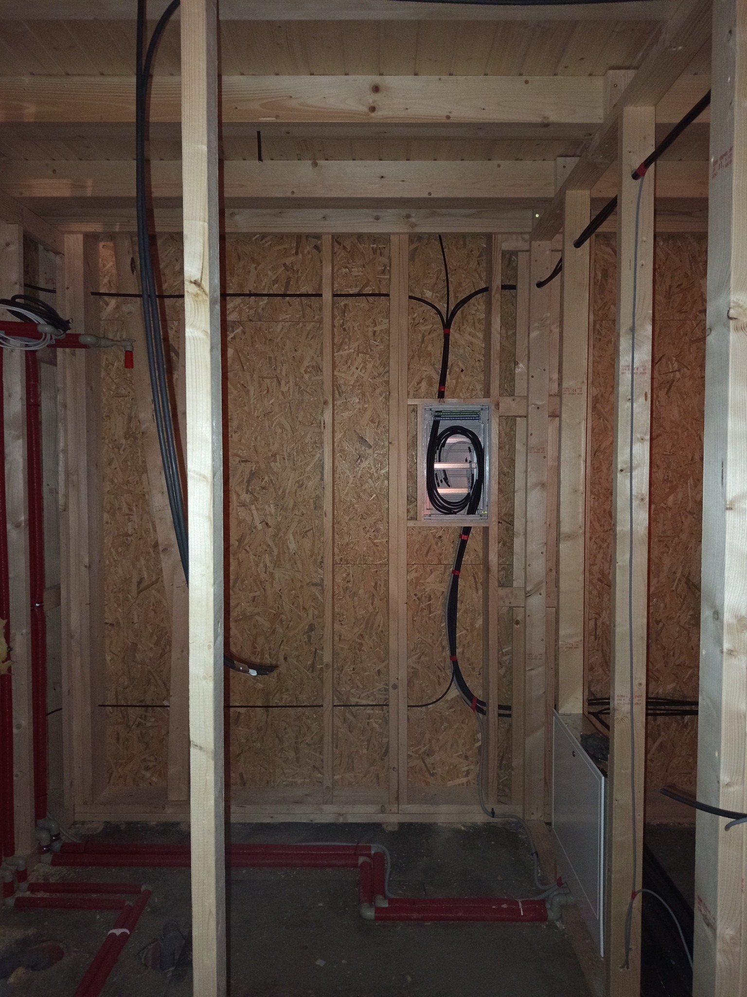 Wnętrze w trakcie budowy: drewniana konstrukcja szkieletowa z widocznymi instalacjami elektrycznymi w puszce i rurami wodnymi w kolorze czerwonym.