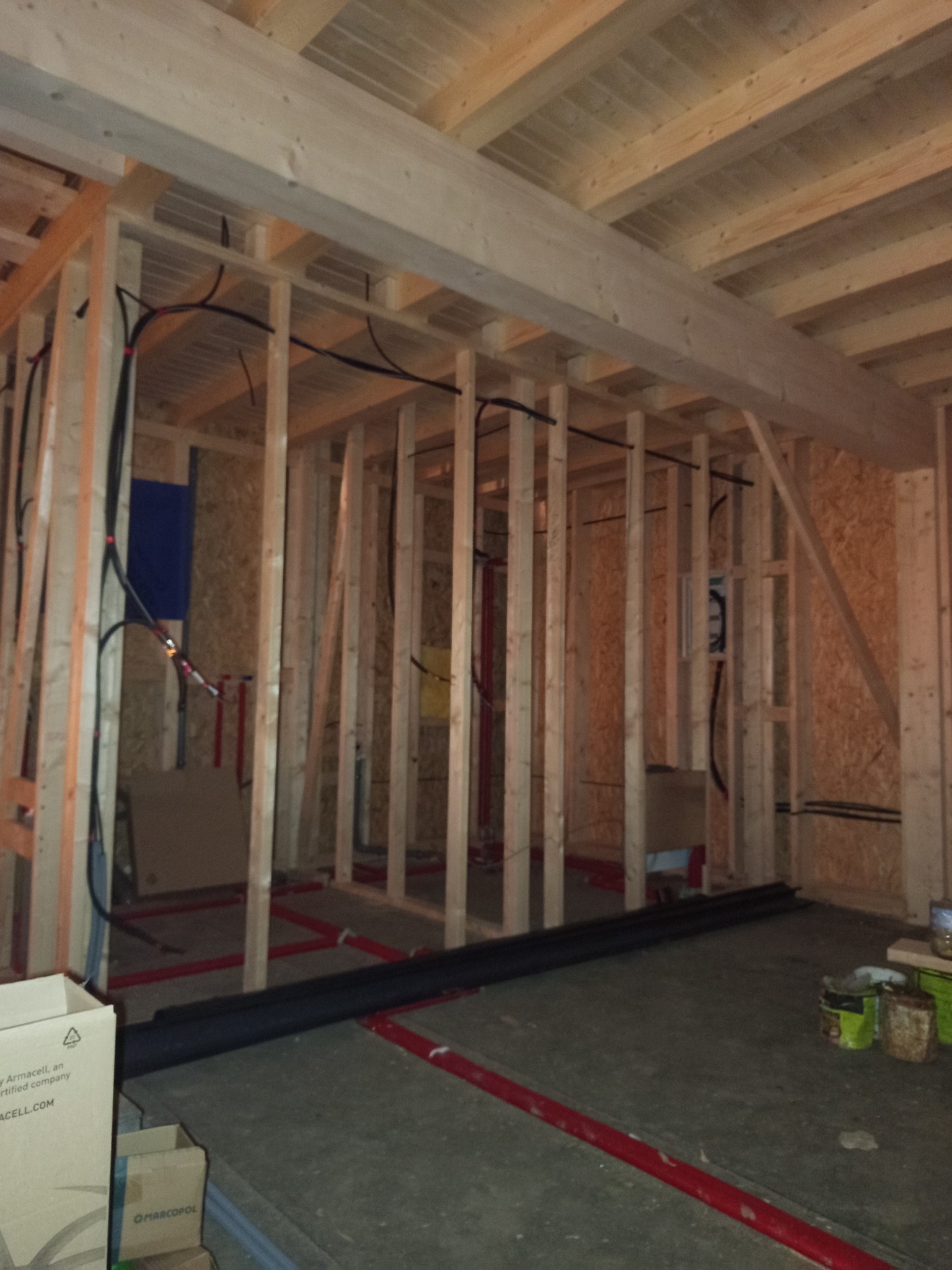 Wnętrze w trakcie rozbudowy: drewniana konstrukcja szkieletowa, instalacje elektryczne w peszlach, rury ogrzewania podłogowego. Widoczne materiały budowlane i narzędzia.