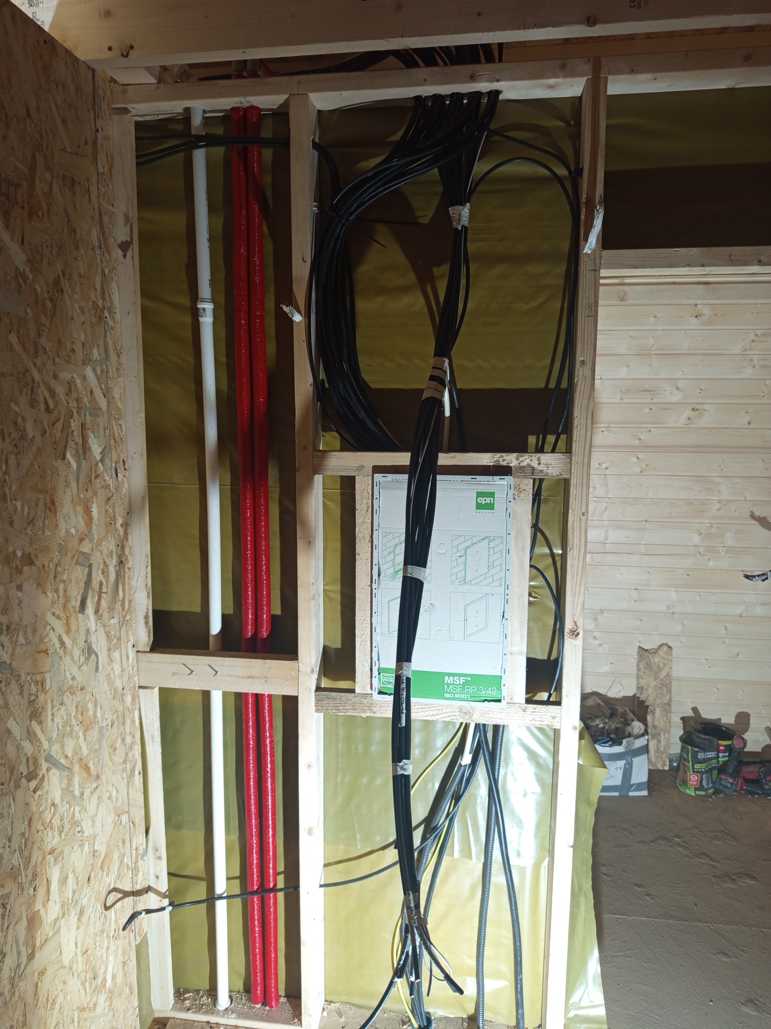 Instalacja elektryczna i rury w drewnianej konstrukcji ściany, widoczne kable w peszlach i puszka instalacyjna. Żółta folia w tle. Prace instalacyjne w trakcie budowy.