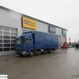 Niebieski samochód ciężarowy zaparkowany przed budynkiem z bramą garażową i żółtym szyldem 'GP Truck Invoice', w tle widoczne inne pojazdy i sprzęt.