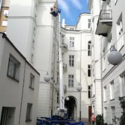 Podnośnik koszowy na dziedzińcu kamienicy z białą elewacją, widoczne pomarańczowe pachołki i osoba w oddali.