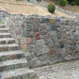 Kamienna ściana oporowa z granitowymi schodami, otoczona zielenią i kostką brukową. Różnorodność kolorów kamieni.